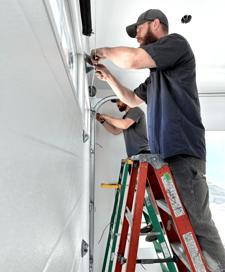 Two men collaboratively repairing a garage door, focused on their task with tools in hand and a determined expression